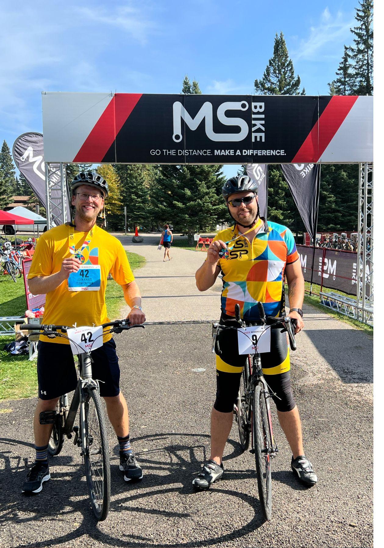 Brennan and a fellow rider standing with their bikes, both wearing bright yellow shirts, black pants, and black bike helmets. They're holding their finisher medals and smiling at the event finish line.